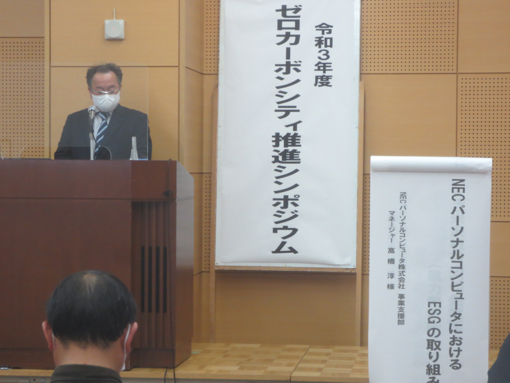 眼鏡をかけマスクをしたNECパーソナルコンピュータ株式会社の男性が演台に立ち話をしている写真