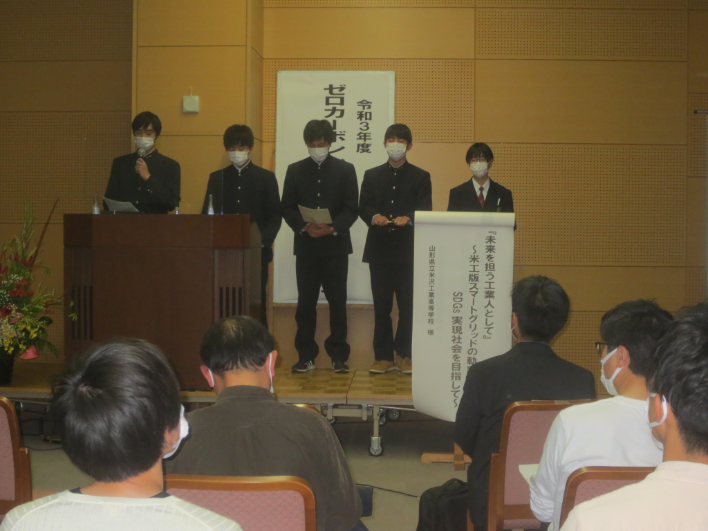 山形県立米沢工業高等学校の男子生徒4名と女子生徒1名が演壇に立ち、左側の男子生徒がマイクを持ち話をしている写真
