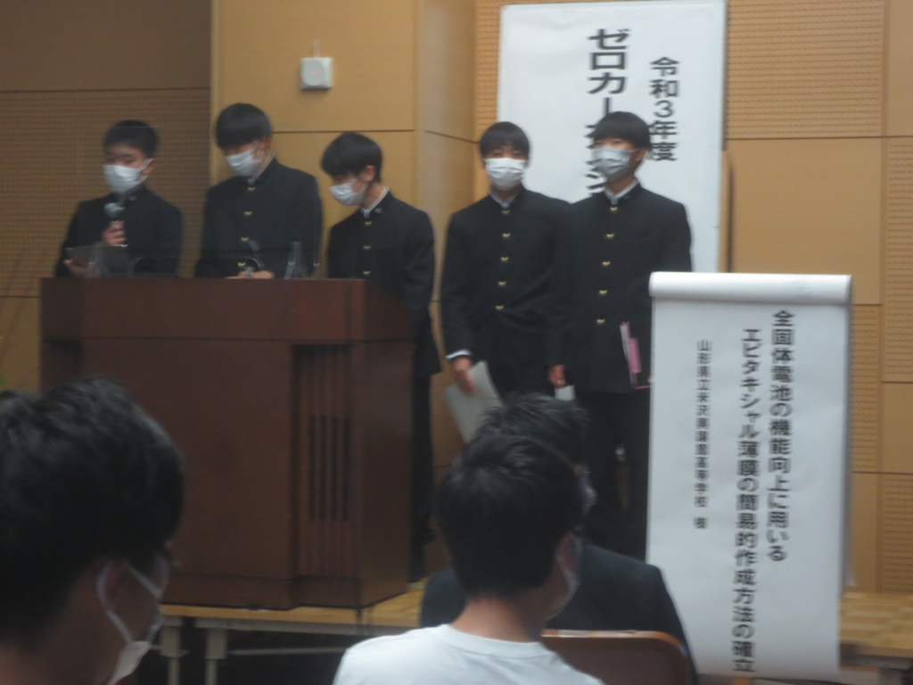 山形県立米沢興譲館高等学校の男子生徒5名が演壇に立ち、左側の男子生徒がマイクを持ち話をしている写真