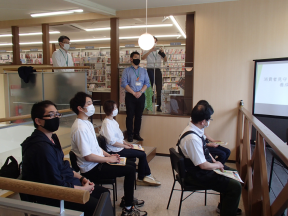 モニターが置かれた部屋で、椅子に座る5名の参加者たちと、奥に立っている関係者たちの写真