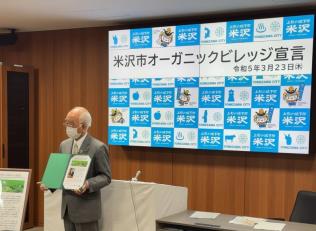 男性が「米沢市オーガニックビレッジ宣言」と書かれたモニターの前で額を両手で持って立っている写真