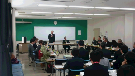 前で男性が立って話をしているのを関係者が座って聞いている懇談会の様子の写真