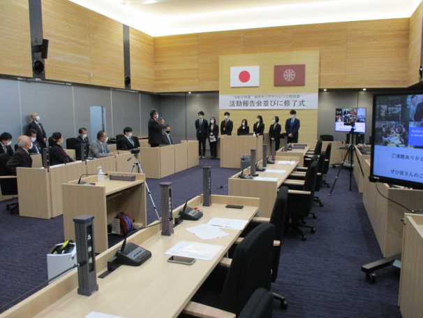 国旗を背に7人スーツを着た男女が立ち、左側に各席にスーツ姿の人達が座りそのうちの1人男性がマイクを持って立っている人達に向かって話している様子の写真