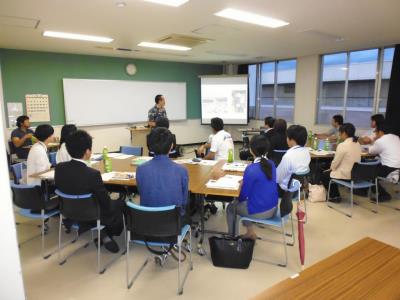 ホワイトボードが設置された広い部屋で、正面に立つ男性の方に椅子を向けて着席した関係者たちが講演を聞いている様子の写真
