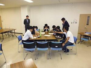 机を取り囲んで、班員が話し合いをしている様子とその後ろで男性2名が話し合いを見ている様子の写真