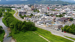 住宅街と川が流れる街の鳥瞰写真。緑豊かな河川敷、道路、家々、ビルなどが写っている。背景には山々が見える。