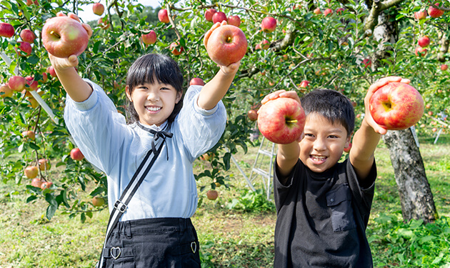 リンゴの木の前で笑顔でリンゴを掲げる女の子と男の子の写真。背景にはたくさんのリンゴが実っている木々が見える。