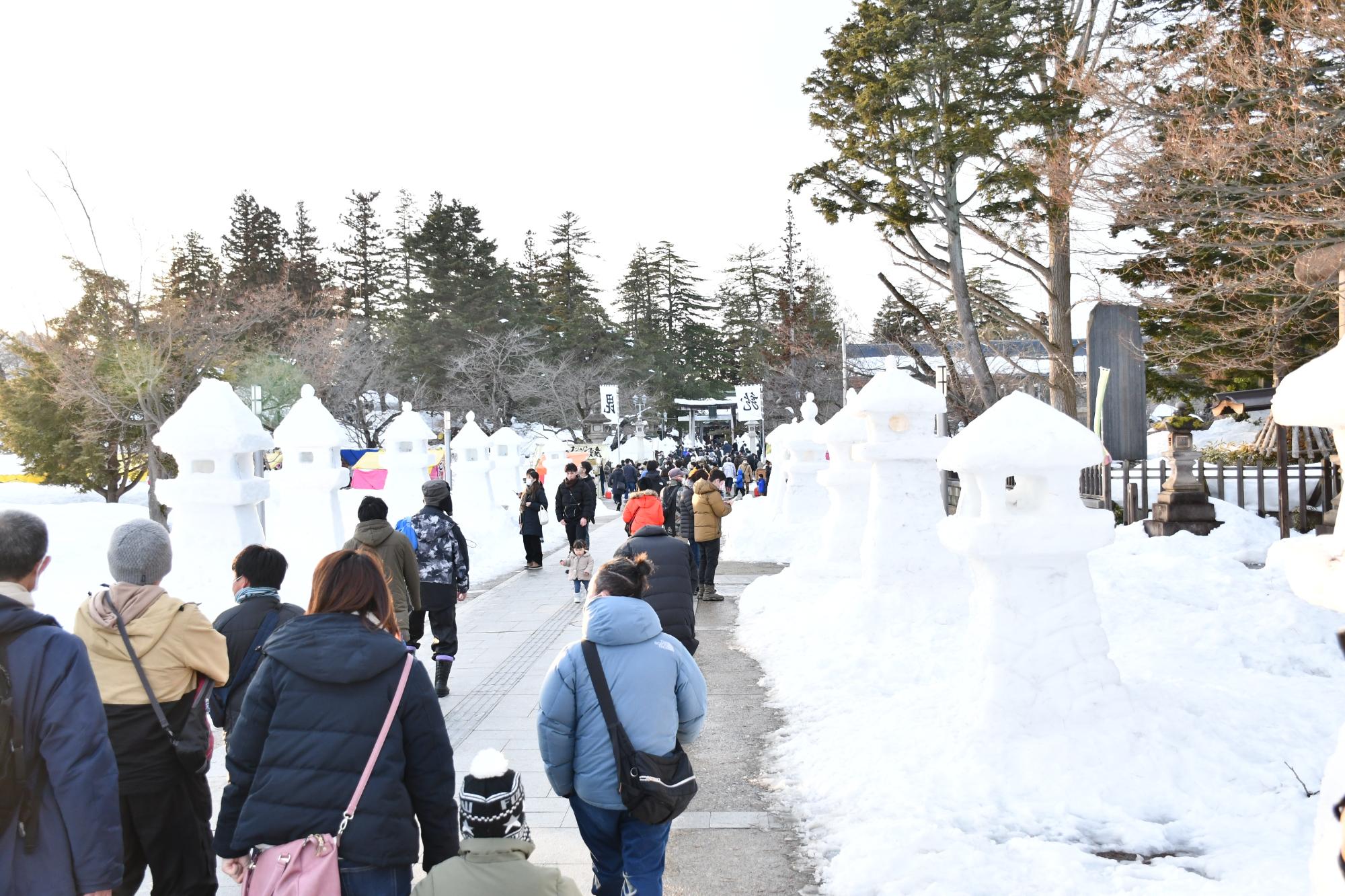 昼間、雪灯籠が並び立つ参道を歩く人々の画像