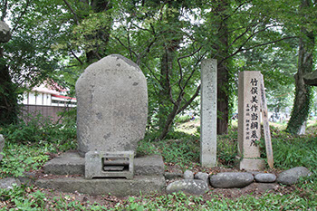 木々に囲まれた中左から大きな俵型の墓石とそれと同じくらいの高さの長方形石造があり、その右に竹俣美作当綱墓所と彫られた石造がある写真