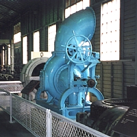 発電所の屋内に設置された水色で上部が筒状になっており、円形のハンドルのようなものがついている発電機とタービンの写真