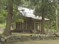手前の石が積み上げられた石堤の上に大きな木が立っており、その奥に見える木造の羽黒神社の外観写真