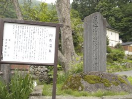 右側に「直江城州公鉄砲鍛造遺跡」と彫られた碑があり、後ろに2本に枝分かれした木が生えており、左手に文章がかかれた白いボードが立ててある直江兼続の鉄砲鍛造遺跡の写真