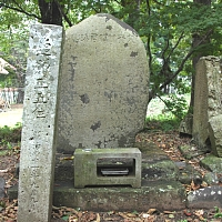 木々を背に中央に草履型の墓石とその手前に長方形の線香入れがあり、左側に長方形の棒型の石が立っている竹俣当綱の墓の外観写真