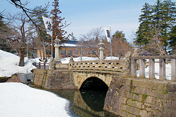 雪の降り積もる場所にアーチ式の石橋が渡っており、水面に映る橋とで眼鏡のように見える舞鶴橋の写真