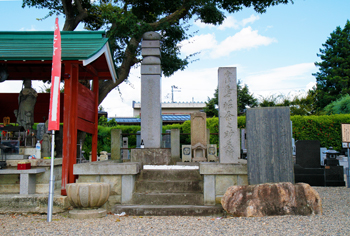 左側に緑色の屋根で赤い木で囲ってある建物の中に仏堂やお供え物があり、その横にある堀粂之助の墓を正面からうつした写真