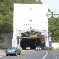 手前から奥に続く道路の先に、鉄筋で作られた入口と西栗子トンネルと書かれた白い壁のトンネルに車が往来している写真