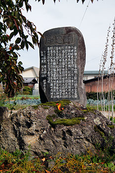 民家の前の畑の中に建っている石碑で、その裏に人の名前がたくさん書かれてある写真