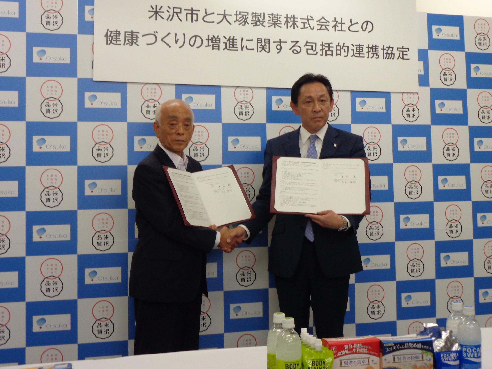 米沢市と大塚製薬株式会社との健康づくりの増進に関する包括的連携協定と書かれた看板を背に手前中央でスーツ姿の2人が片手で握手をし、もう片方の手にそれぞれ協定書を持ちカメラに向けている様子の写真