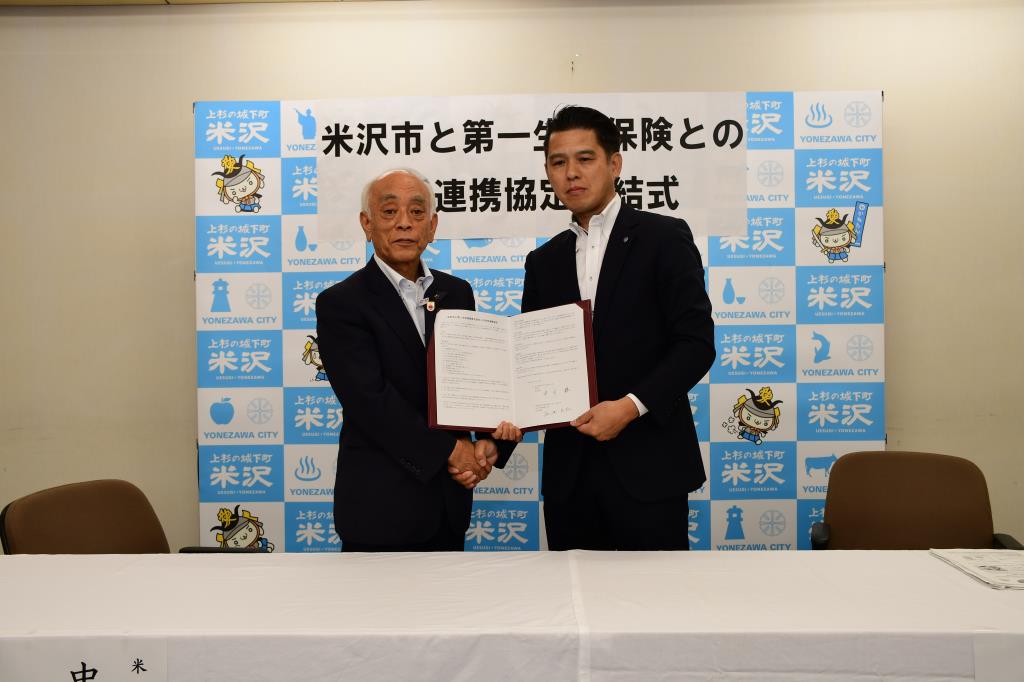 米沢市と第一生命保険株式会社との包括連携協定締結式と書かれたボードを背に中川市長と谷池氏が書類を持ち此方に見せている写真