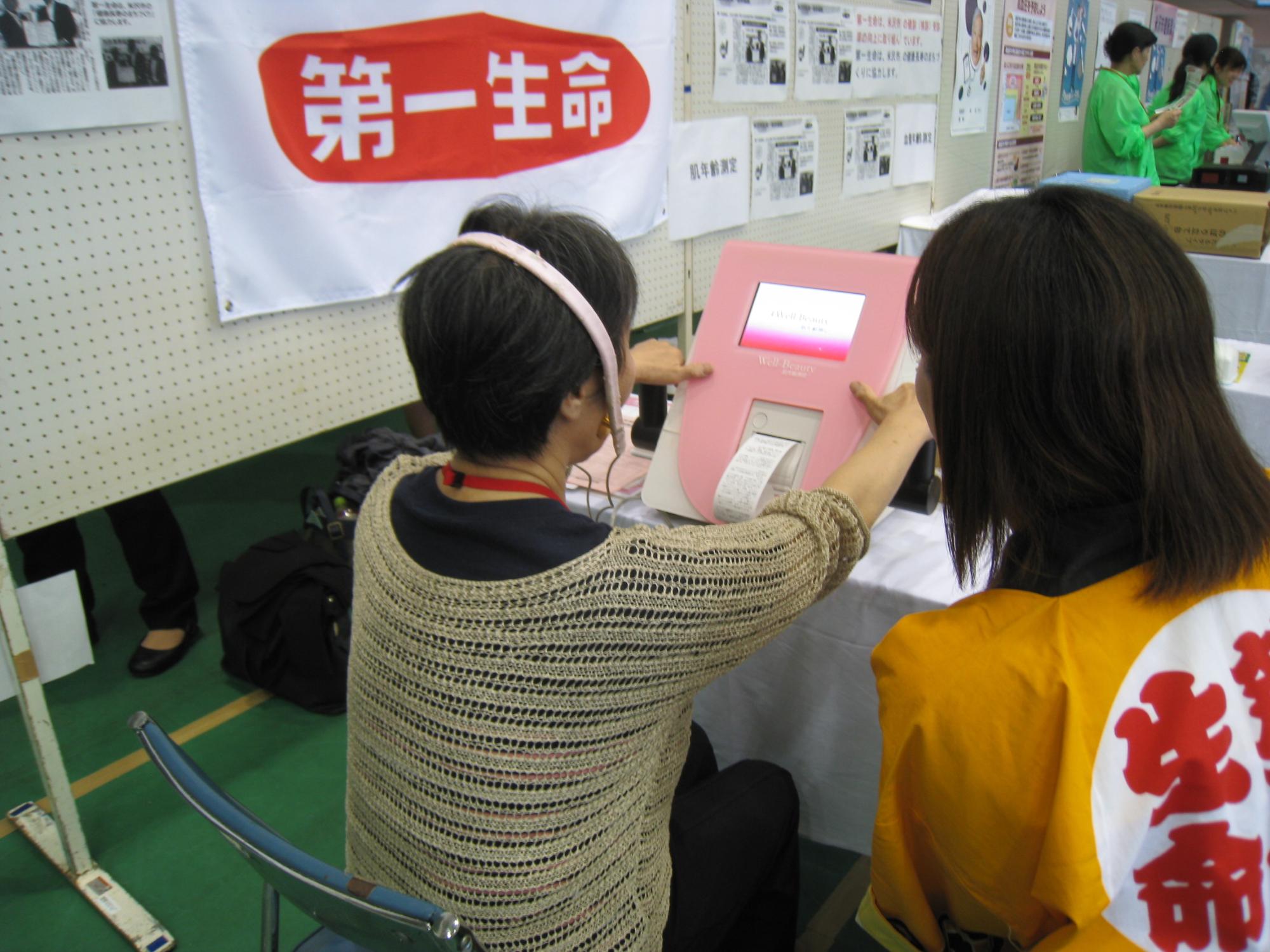 手前にいるオレンジ色のはっぴを着た女性が肌年齢と血管年齢を測定出来る機械を使っている女性と一緒にいる様子の写真