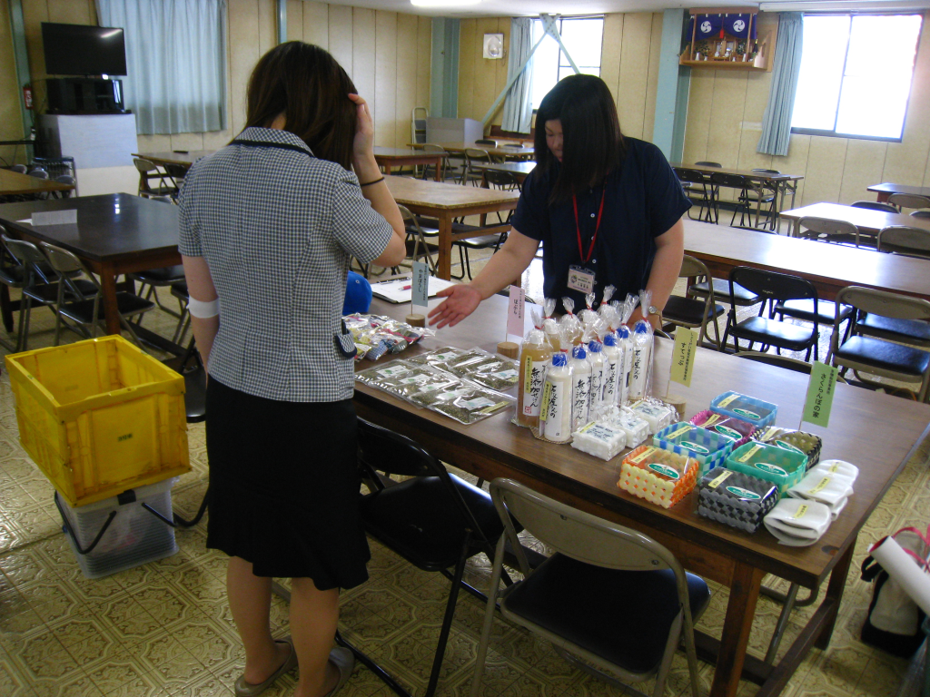 茶色いテーブルの上に置かれた石鹸やわらじ、編んで作られたカゴなどの記念品について説明をするスタッフの女性と、話を聞く制服を着た女性の写真