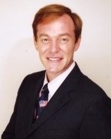 紺のネクタイに黒いスーツを着て微笑むブロンドの髪を軽く七三分けにしている男性ダニエル・カールさんの写真