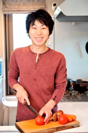 赤褐色の長袖を着た男性がキッチンでまな板の上のトマトを包丁で切ろうとしている相田幸二さんの写真