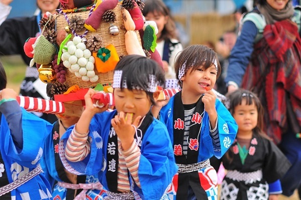 青い法被姿の子どもたちが、松ぼっくり、サツマイモ、ブドウ、大根、トマトなどのオーナメントが飾られた神輿を担いでいる様子の写真
