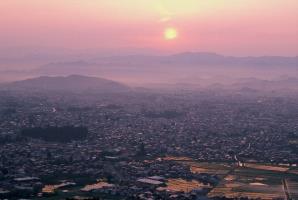 田園や街並み、山並みが広がり、正面から日の出が昇っている様子を撮った風景写真