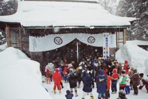 雪が降り積もる中、白地に二羽飛び雀の家紋が入った神社幕の奥から袴姿の人々が両手を掲げた防寒着姿の参加者達に向け豆を巻いている様子の写真