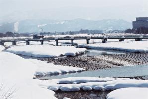 手前に、両岸に雪が降り積もった穏やかな川が流れる松川と、一台のトラックが走る松川に架かる住之江橋があり、奥には吾妻山を望む写真