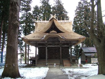 大きな樹木が周りに生えている場所で、末広がりで立派な茅葺屋根の羽黒神社本殿が建っている写真
