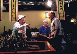 値段が書かれた紙や電球が下がった店内で、帽子を被った人が商品を持って男性に声をかけている様子の写真