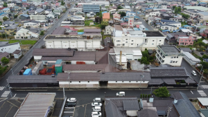 黒や褐色の大きな屋根をもち横に長く続く建物である小嶋総本店とその他の建物が並んで立つ全景を上空から撮影した写真