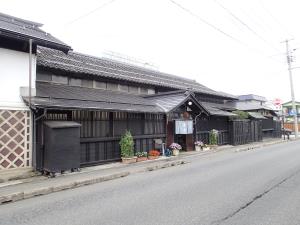 横に長く続く日本家屋であり、入り口付近に花が咲いた植木鉢が置いてある小嶋総本店店舗兼主屋を道路を挟んだ斜め向かい側から撮影した写真