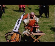鎧を身に着けた人たちが芝生の広がる場所に集まっており、1名の兜を脱いだ人が台の前に立って儀式を行っている様子の写真