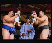 2名の力士のかたが向き合っており、それぞれ抱き上げた赤ちゃんを向かい合わせるようにしている様子の写真