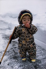 フード付きのオールインワンの洋服を着た幼児が、手に掴んだ雪を口に運んでいる写真