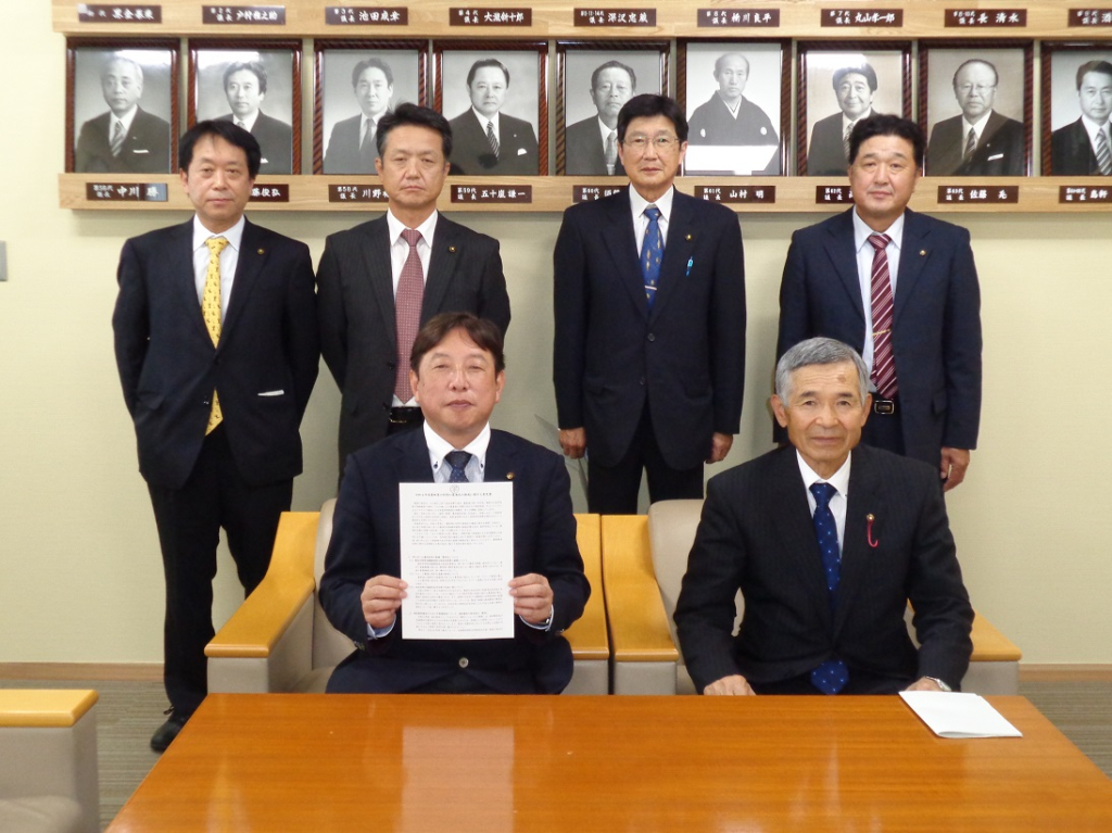 歴代議長の白黒写真が飾られている壁の前で、関係者の男性4人が立って並び、前列に意見書を持った市議会議長と男性が座り記念撮影をしている写真