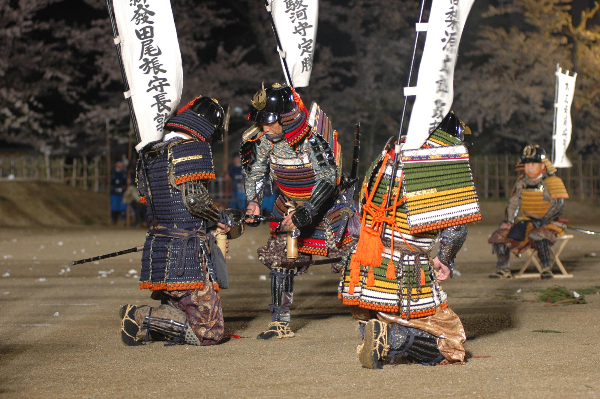 甲冑を着た武者が膝をついて控える武者が持っている入れ物へ柄杓に入った水を注いでいる儀式の一幕の写真