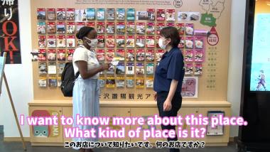 観光案内所で、外国人女性がパンフレットを手に持ち職員に尋ねている写真で、お店について質問している英文と日本語訳がテロップで表記されている写真（YouTube）