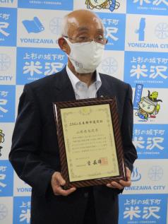 米沢市のロゴが入った水色と白を基調とするバックボードの前に市長が立ち、胸の部分に賞状を持っている写真