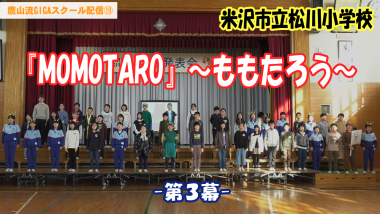 体育館の舞台に40人ほどの小学生が横並びで正面を向いて立っており、中央に「MOMOTARO」～ももたろう～第3幕と大きくテキストが入っている写真（YouTube）