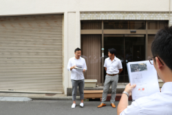 シャッターやガラス戸が閉められた道路沿いに2名の関係者が並んで立っており、道路の向かい側に立つ1名の男性が手に持つ資料を眺めている様子の写真