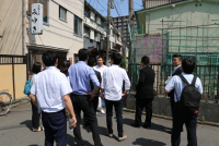 周りに建物が建ち並ぶ道路上に関係者の方々が集まって話をしている様子の写真