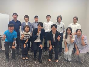 民間交流団体ドットミートの参加者男女が2列になり後ろに6人立ち、前の6人は中腰で笑顔でピースサインをしている集合写真