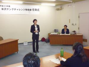 黒いスーツ姿の女性が前に立ち、白い紙を手に持ちながら笑顔で参加者の方を向いて自己紹介をしている様子の写真