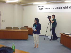 ジャケットを着た女性が前に立ち、紙を両手に持ちながら参加者の前で自己紹介をしている様子の写真