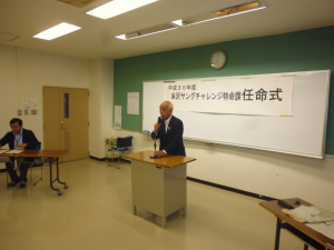 正面に「米沢ヤングチャレンジ特命課任命式」と書かれた紙が設置された会場で、市長がマイクに手を添えながら話をしている様子の写真