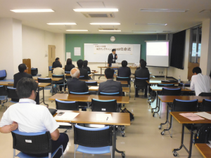 等間隔に設置されたテーブルに2脚ずつの椅子が用意された会場で、着席した関係者たちが正面に立つ男性の方を見ている様子の写真
